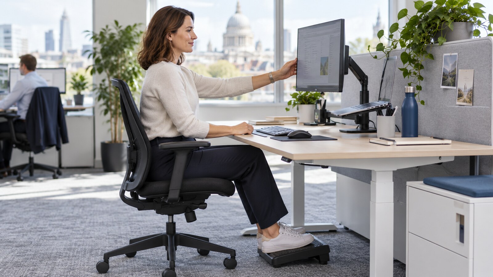 Office worker adjusting a monitor at an ergonomic workstation with a supportive chair, footrest, separate keyboard and mouse, and laptop on a stand