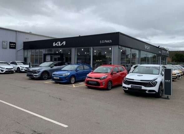 Exterior view of a busy Kia Motors car dealership with vehicles parked outside and a mechanic working on-site, under clear skies.