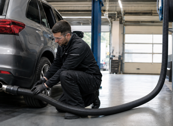 Vehicle technician connecting a tailpipe exhaust extraction hose to a diesel vehicle in a workshop